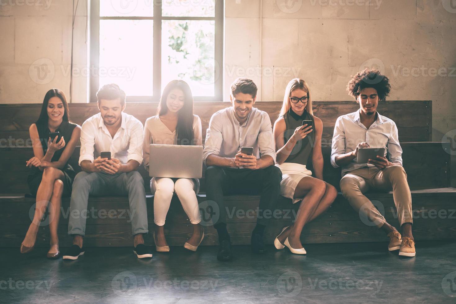 estudantes modernos. vista frontal de jovens usando diferentes gadgets enquanto estão sentados próximos uns dos outros em uma fileira foto