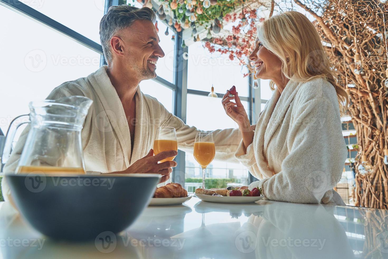 lindo casal maduro em roupões de banho tomando café da manhã juntos enquanto passa o tempo na cozinha doméstica foto