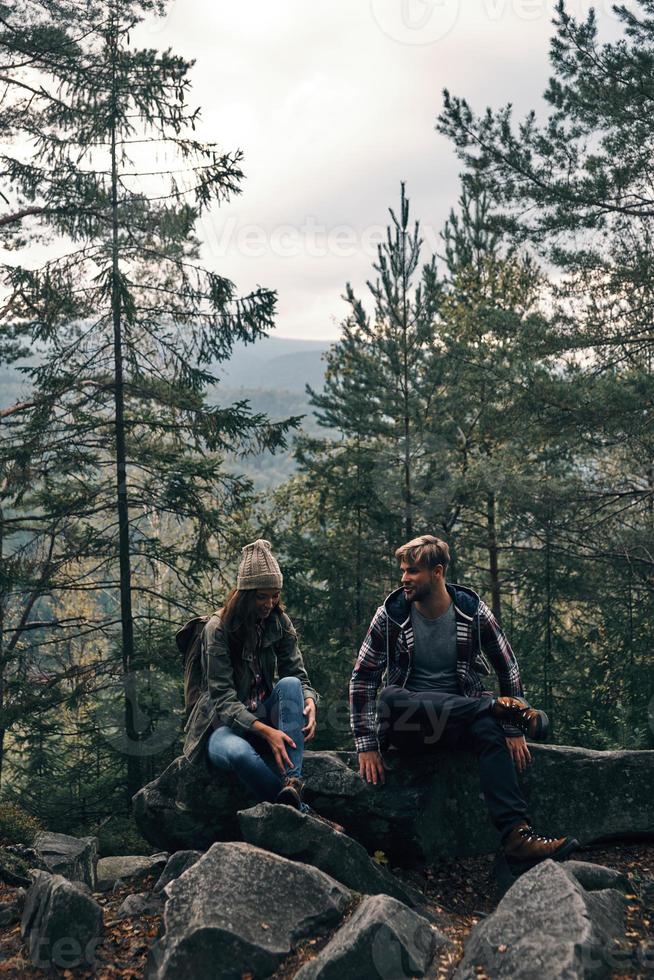 hora de descansar. lindo casal jovem sentado nas rochas e sorrindo enquanto caminhava juntos na floresta foto