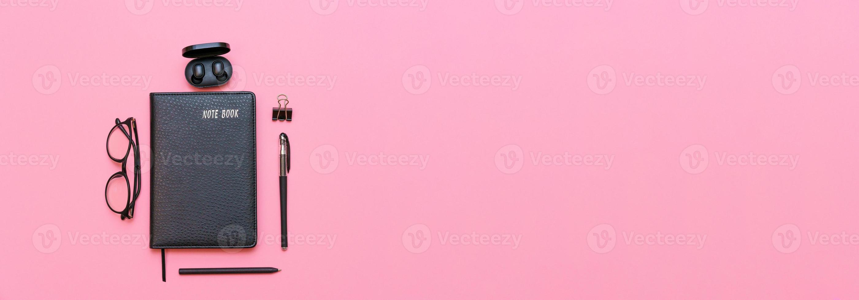 caderno preto com óculos e uma caneta, fones de ouvido sem fio em um fundo rosa. foto