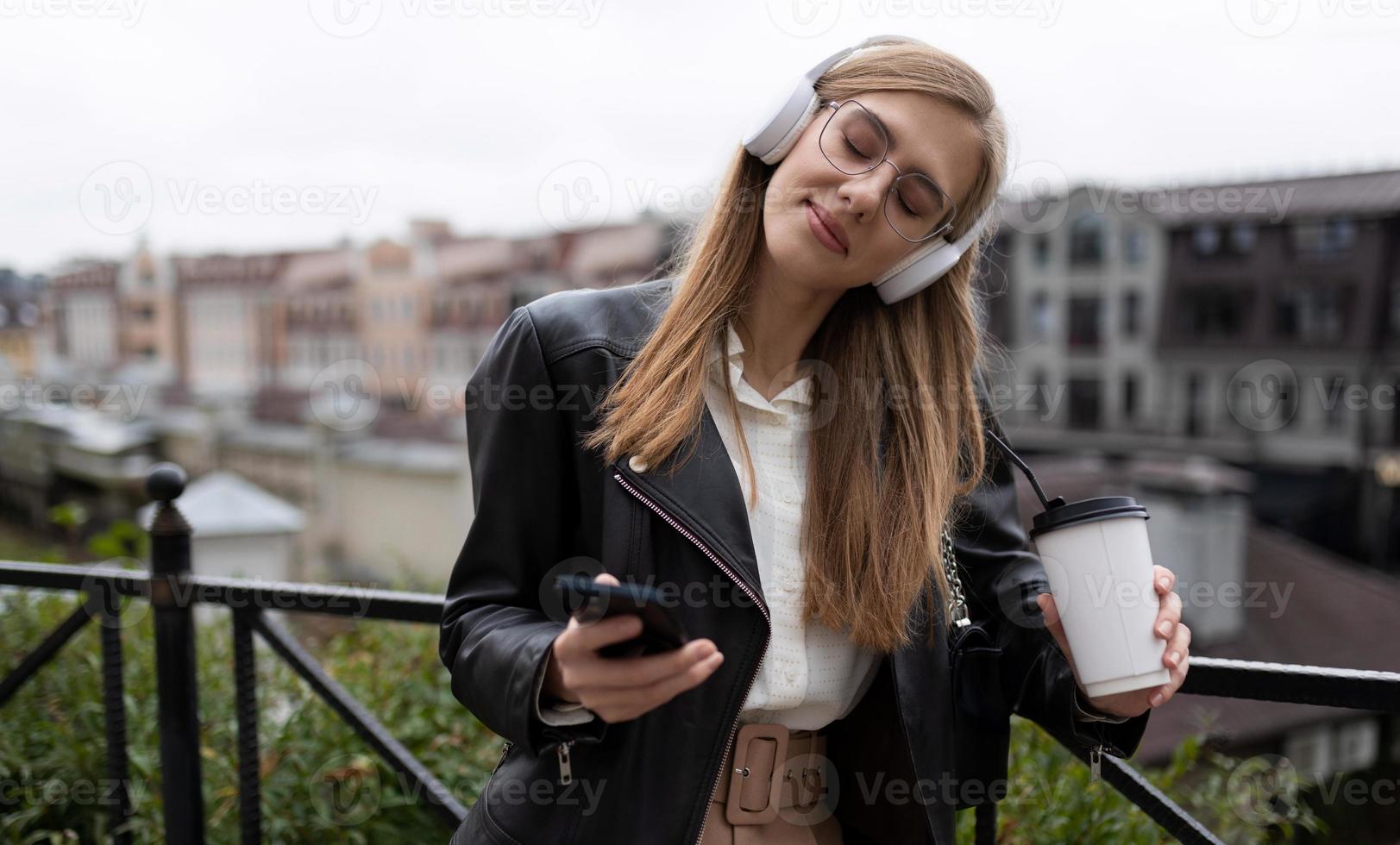 jovem loira para as mãos do celular e um copo de café ouve música em fones de ouvido fechando os olhos com prazer no fundo da paisagem urbana foto