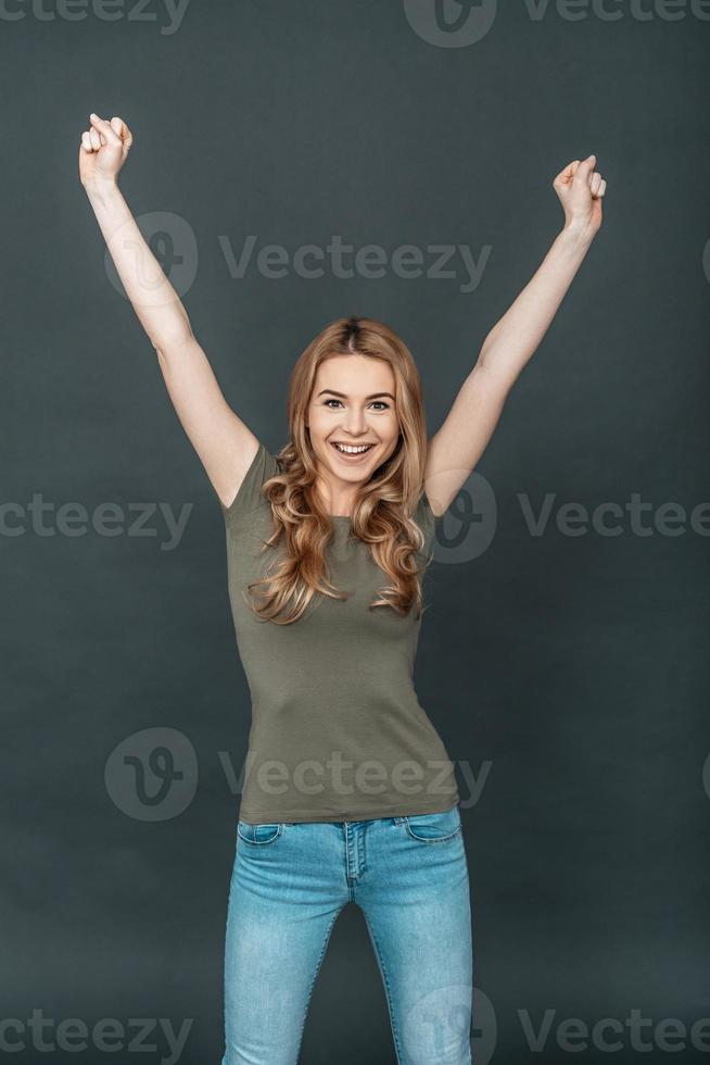 ela é uma jovem feliz vencedora com cabelos loiros em roupas casuais, mantendo os braços levantados e olhando para a câmera em pé contra um fundo cinza foto