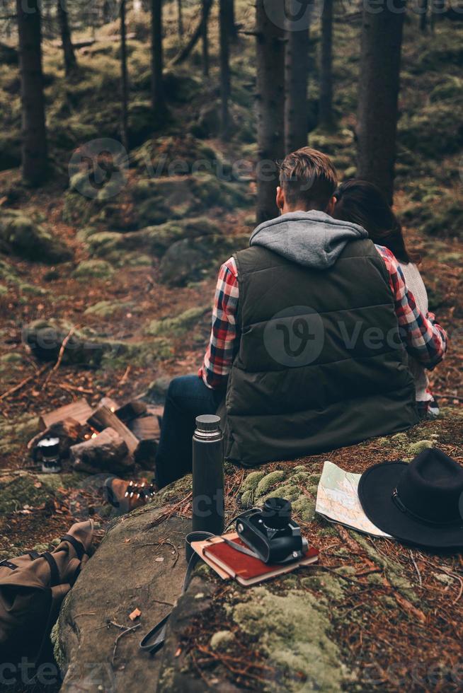 sentindo-se aconchegados juntos. vista traseira do jovem casal aquecendo junto à fogueira enquanto está sentado na floresta foto