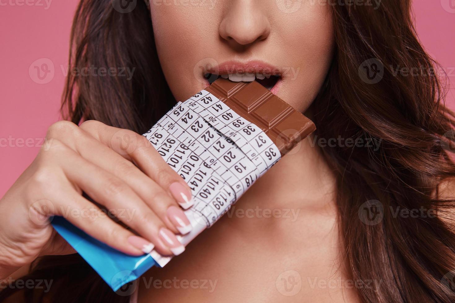 comida proibida. close-up de jovem comendo chocolate embrulhado em fita métrica em pé contra um fundo rosa foto