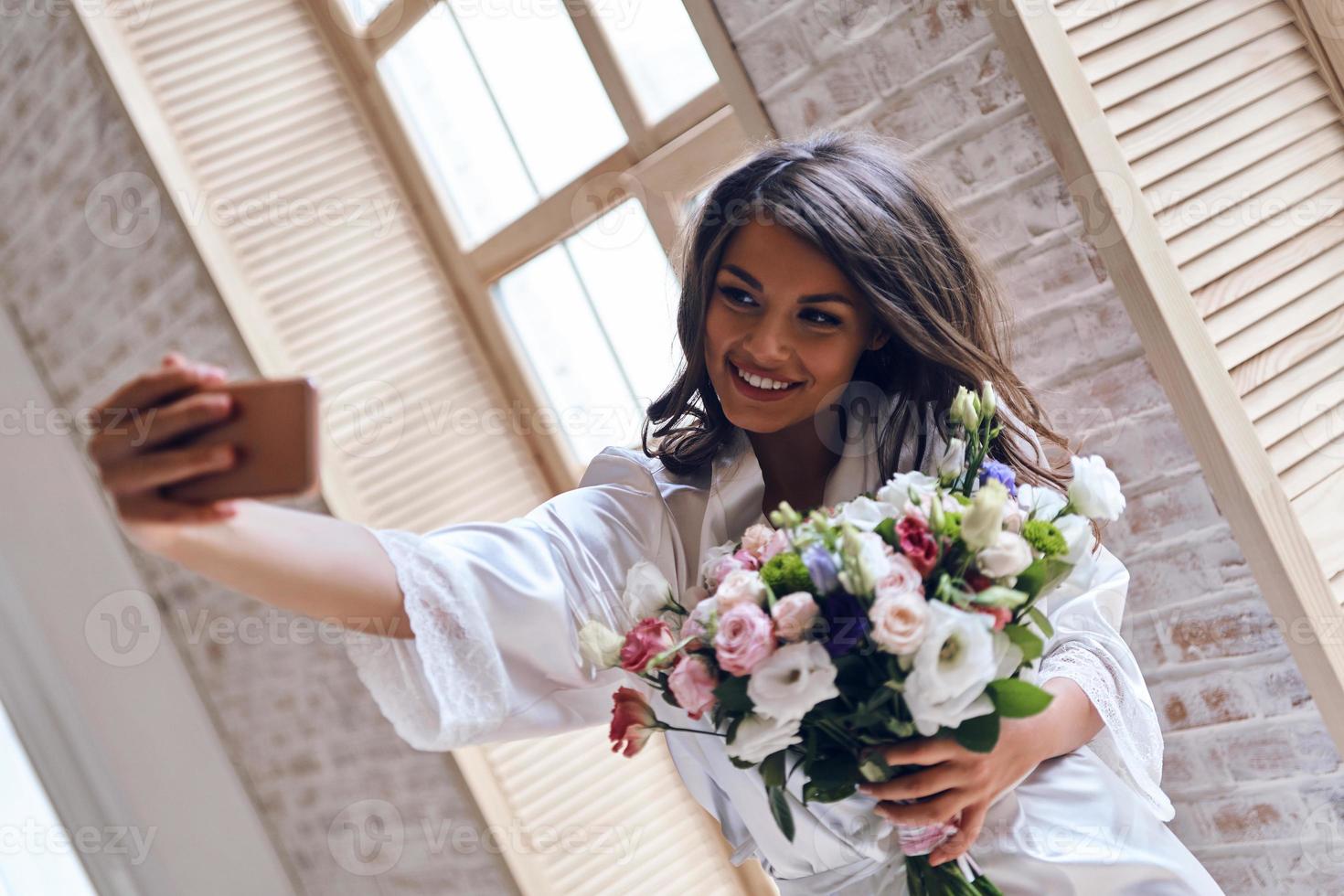 capturando momento feliz. mulher jovem e atraente em roupão de seda segurando um buquê e sorrindo enquanto toma selfie perto da janela foto