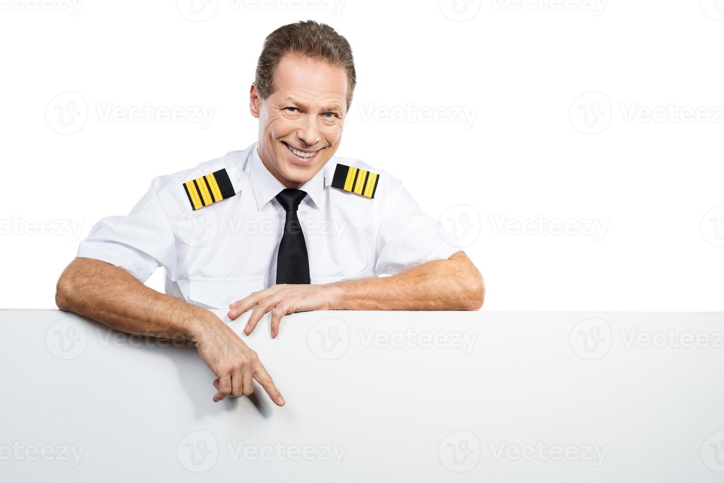 basta olhar para isso. piloto masculino confiante em uniforme, inclinando-se para o espaço da cópia e apontando-o com um sorriso em pé contra um fundo branco foto