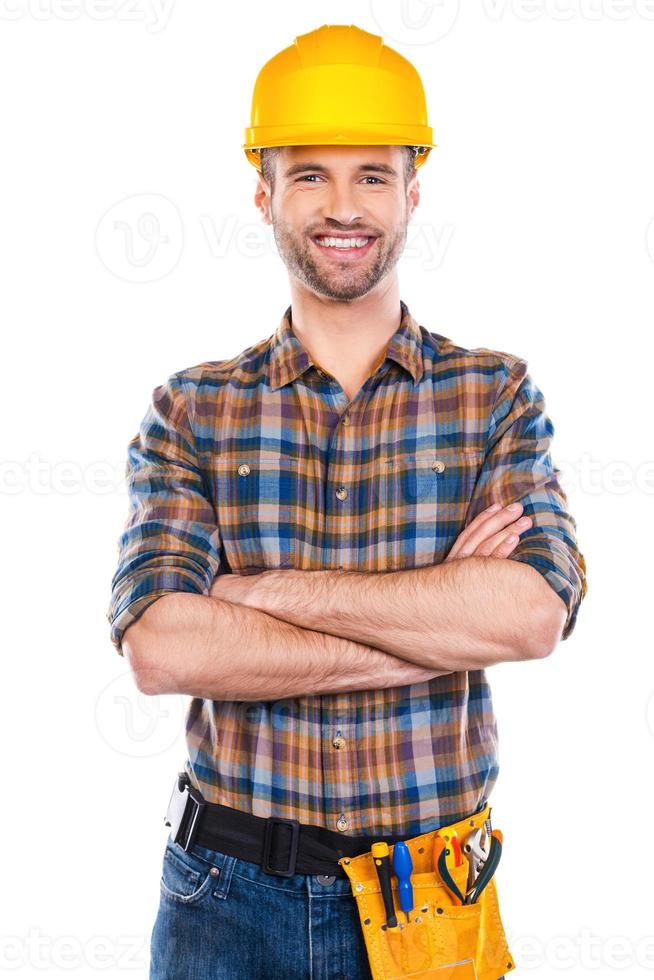 reparador confiante. confiante jovem carpinteiro masculino no capacete de segurança, mantendo os braços cruzados e sorrindo em pé contra um fundo branco foto