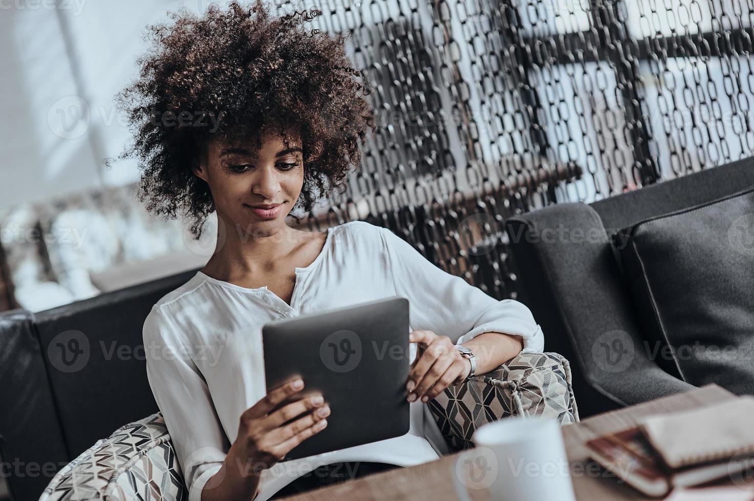 examinando seu novo tablet digital. bela jovem africana em roupas casuais inteligentes trabalhando usando tablet digital enquanto está sentado no café foto
