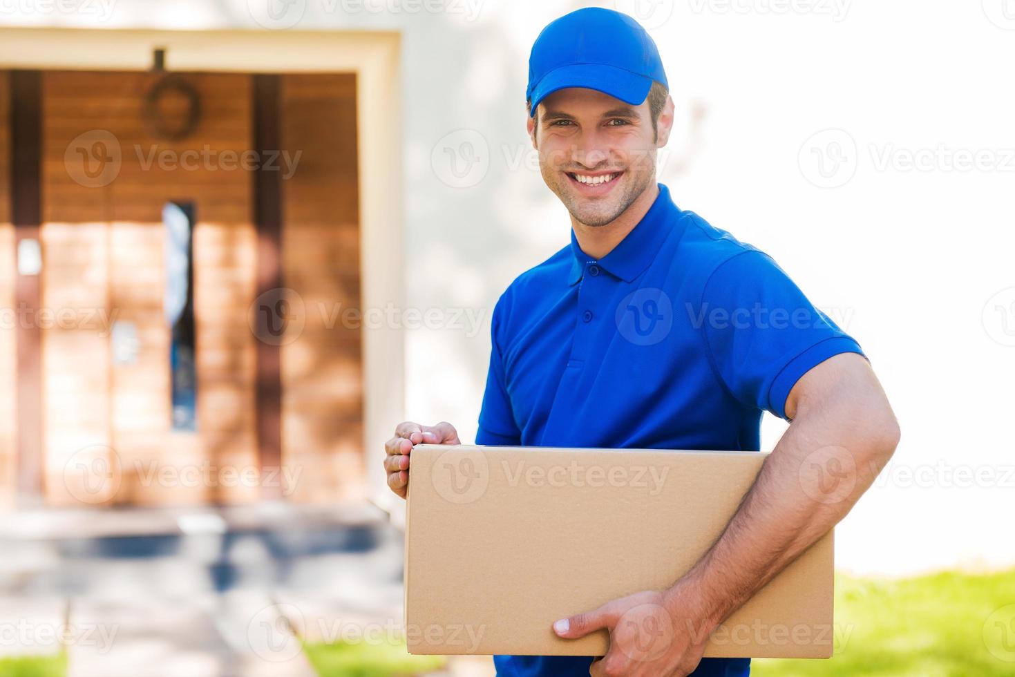 sempre a tempo. alegre jovem entregador segurando uma caixa de papelão em pé contra a casa residencial foto