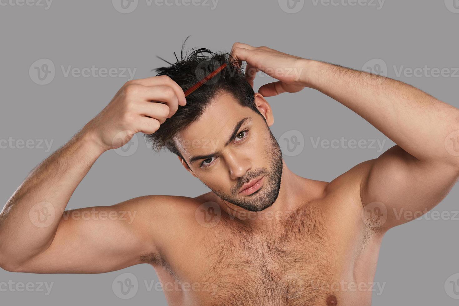 prestando atenção em cada detalhe. jovem bonito penteando o cabelo em pé contra um fundo cinza foto