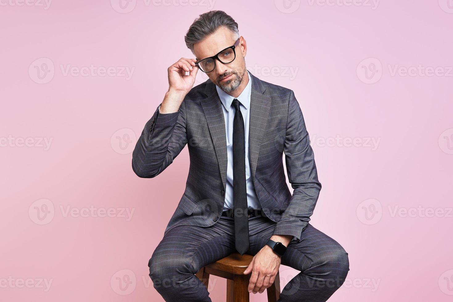 homem maduro bonito em trajes formais, ajustando os óculos enquanto está sentado contra um fundo rosa foto