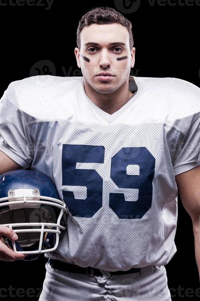 configurado para o jogo. jogador de futebol americano olhando para a câmera e segurando o capacete na mão em pé contra um fundo preto foto