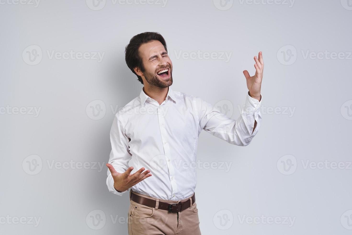 jovem feliz na camisa branca, gesticulando e mantendo os olhos fechados em pé contra um fundo cinza foto