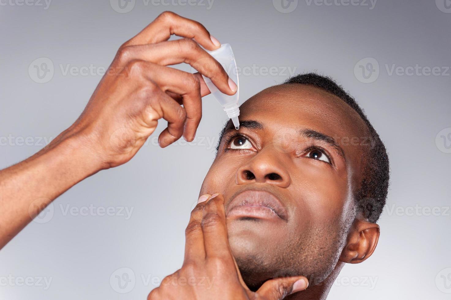 olho pingando do homem. jovem africano aplicando colírio em pé contra um fundo cinza foto