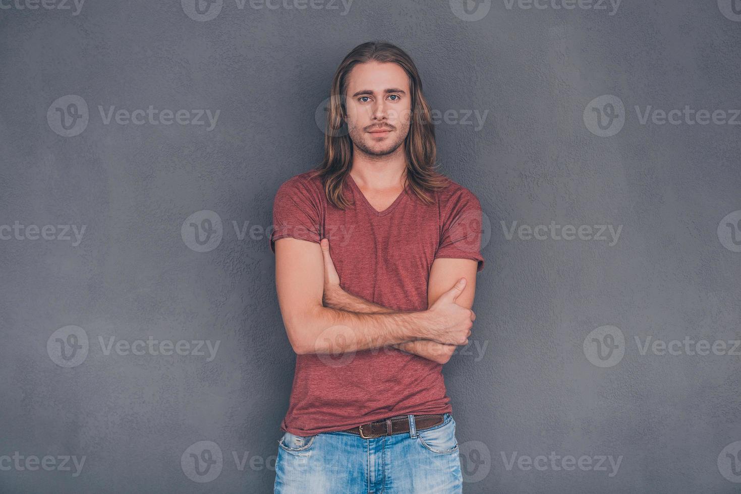 confiante e bonito. jovem bonito com roupa casual, olhando para a câmera e mantendo os braços cruzados em pé contra um fundo cinza foto