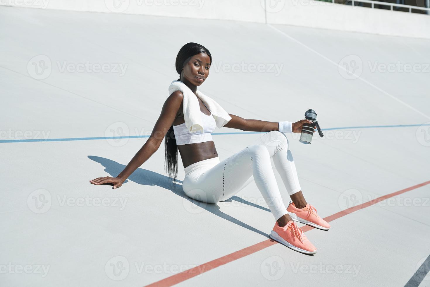bela jovem africana em roupas esportivas, segurando a garrafa com água enquanto está sentado na pista de esportes ao ar livre foto