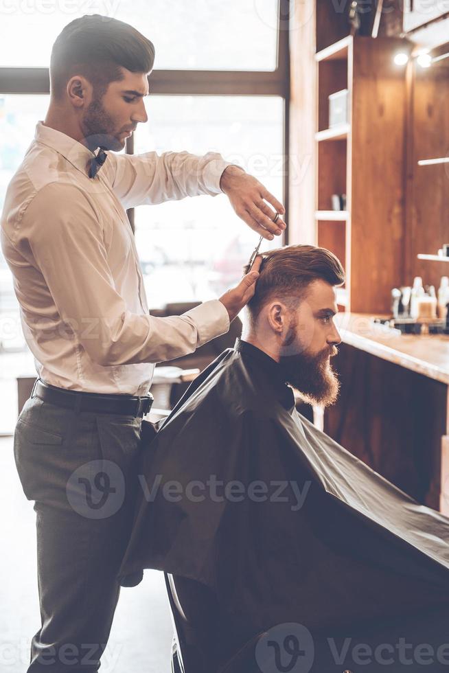 barbeiro no trabalho. Vista lateral do jovem barbudo cortando o cabelo pelo cabeleireiro enquanto está sentado na cadeira na barbearia foto