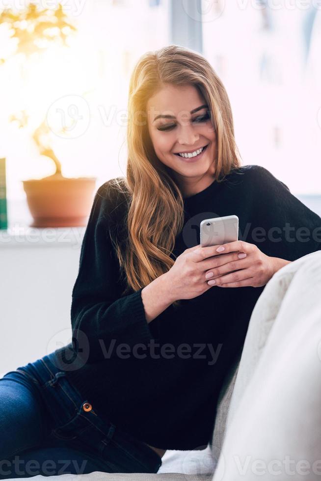 você tem uma nova mensagem linda jovem usando smartphone com sorriso enquanto está sentado no sofá em casa foto