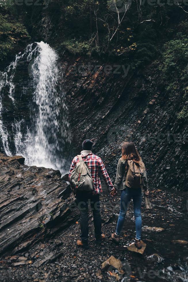 no lugar incrível. vista traseira de comprimento total do jovem casal de mãos dadas em pé perto da cachoeira foto