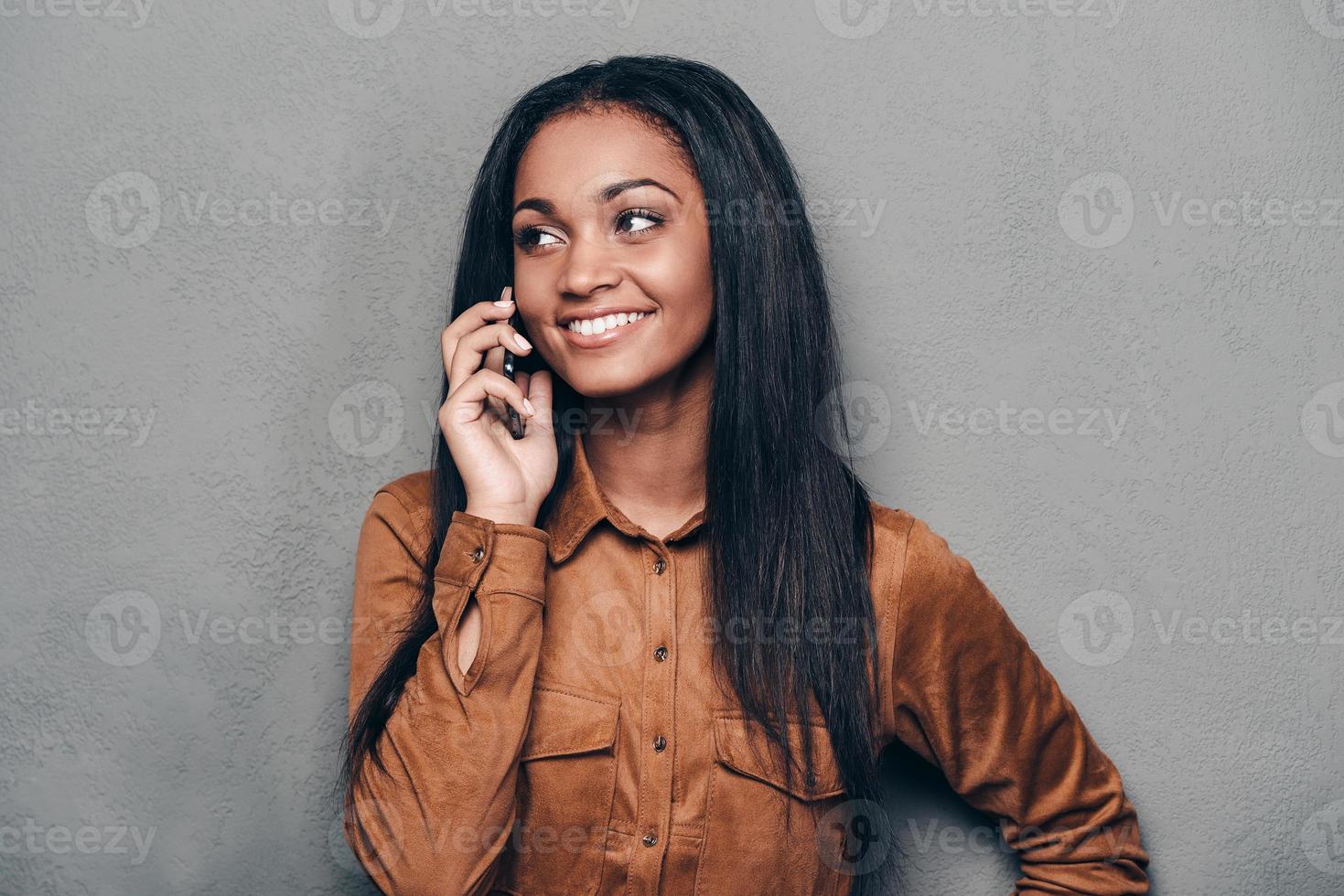 boas notícias. bela jovem africana falando no celular e desviar o olhar em pé contra um fundo cinza foto