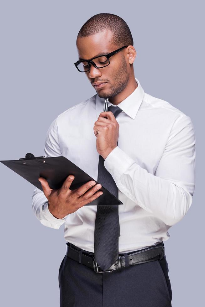 examinar o contrato antes de assinar. pensativo jovem africano de camisa e gravata segurando a prancheta e tocando seu queixo com caneta em pé contra um fundo cinza foto