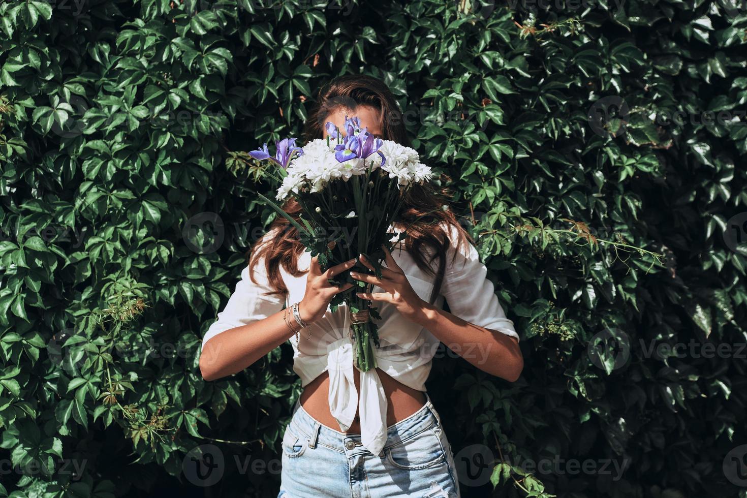 apenas sendo ela mesma. close-up de mulher jovem e atraente em roupas casuais, cobrindo o rosto com um ramo de flores em pé na frente da hera ao ar livre foto