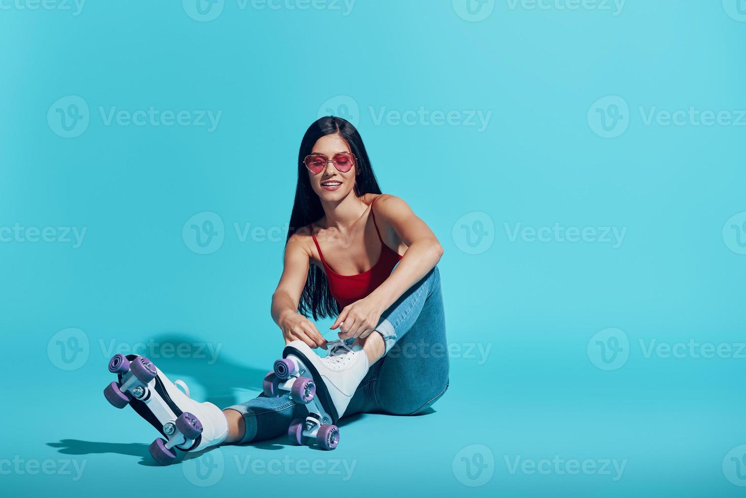 mulher jovem e atraente amarrando o patins e sorrindo enquanto está sentado contra um fundo azul foto