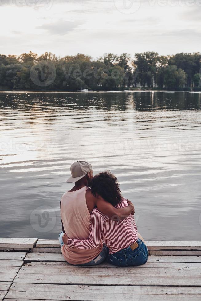 compartilhando amor. vista traseira do jovem casal abraçando enquanto está sentado no cais foto