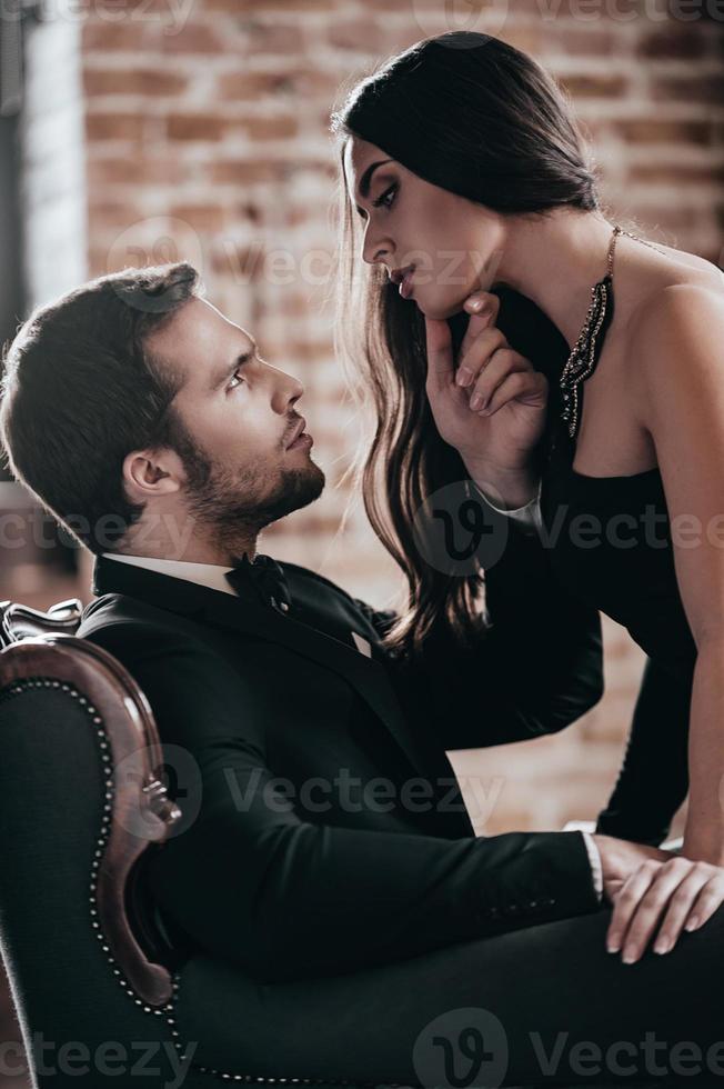 casal sensual. mulher jovem e bonita em vestido de cocktail, inclinando-se para o namorado sentado na cadeira e segurando a mão no queixo dela enquanto se olham no interior do loft foto