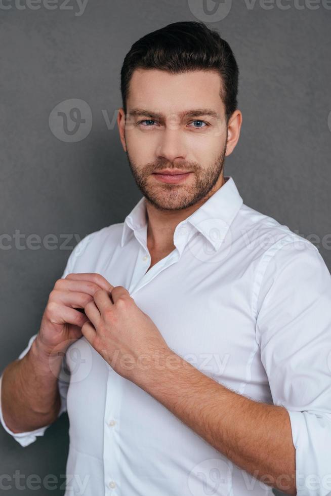 encantador bonito. jovem bonito abotoando sua camisa branca e olhando em pé contra um fundo cinza foto