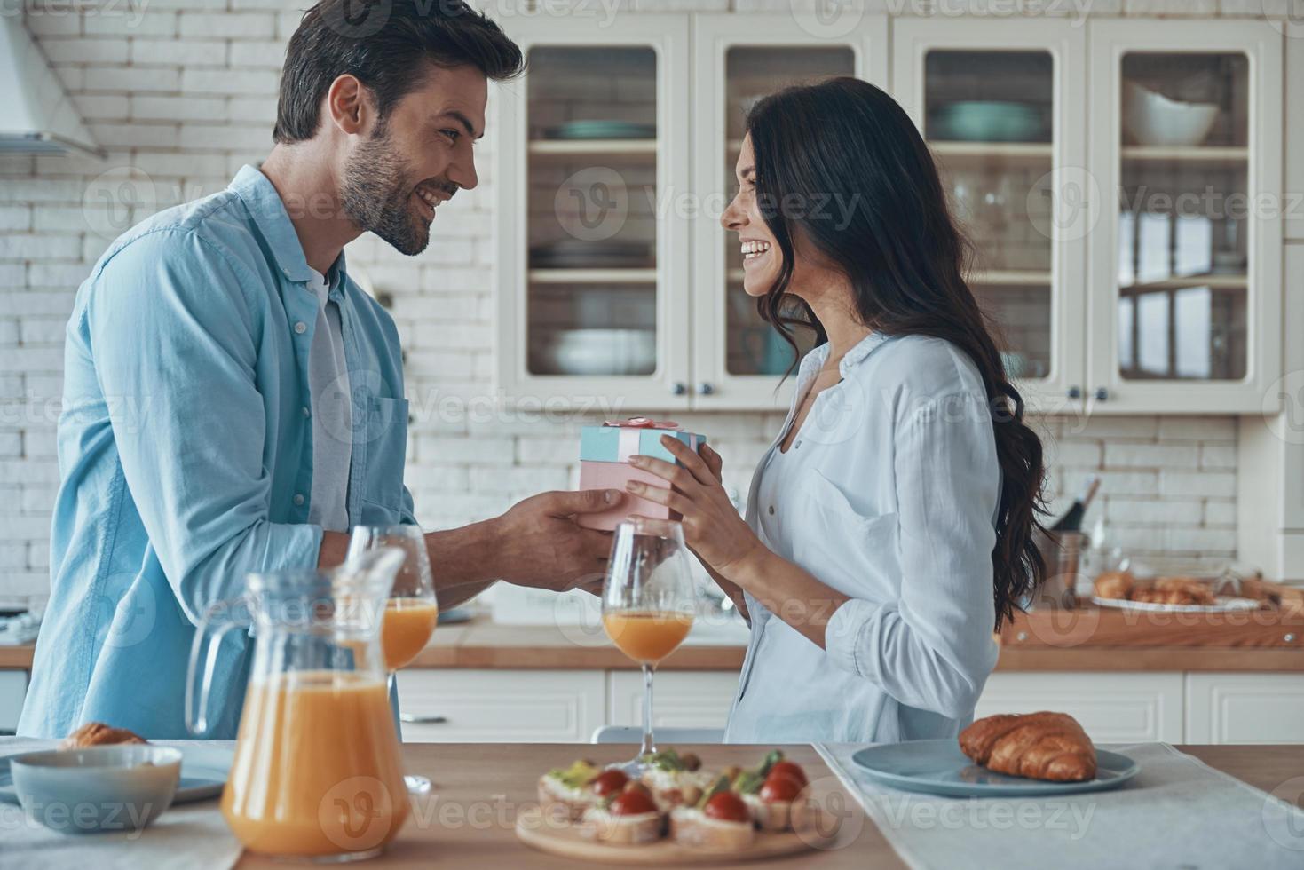 jovem bonito dando uma caixa de presente para sua namorada enquanto toma café da manhã na cozinha doméstica juntos foto