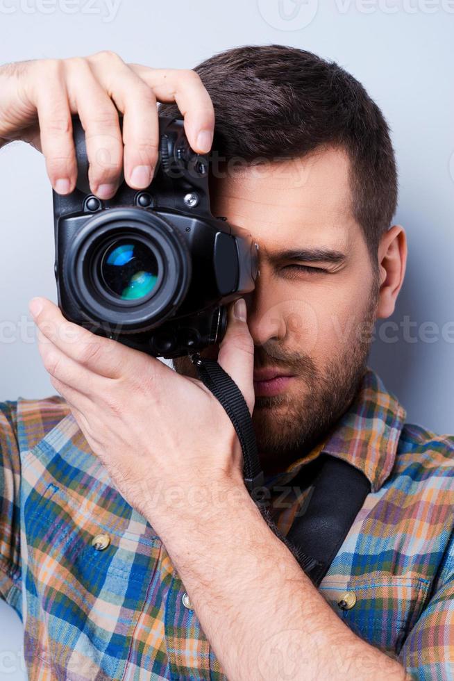 fotógrafo sério. retrato de jovem confiante na camisa segurando a câmera na frente de seu rosto em pé contra um fundo cinza foto