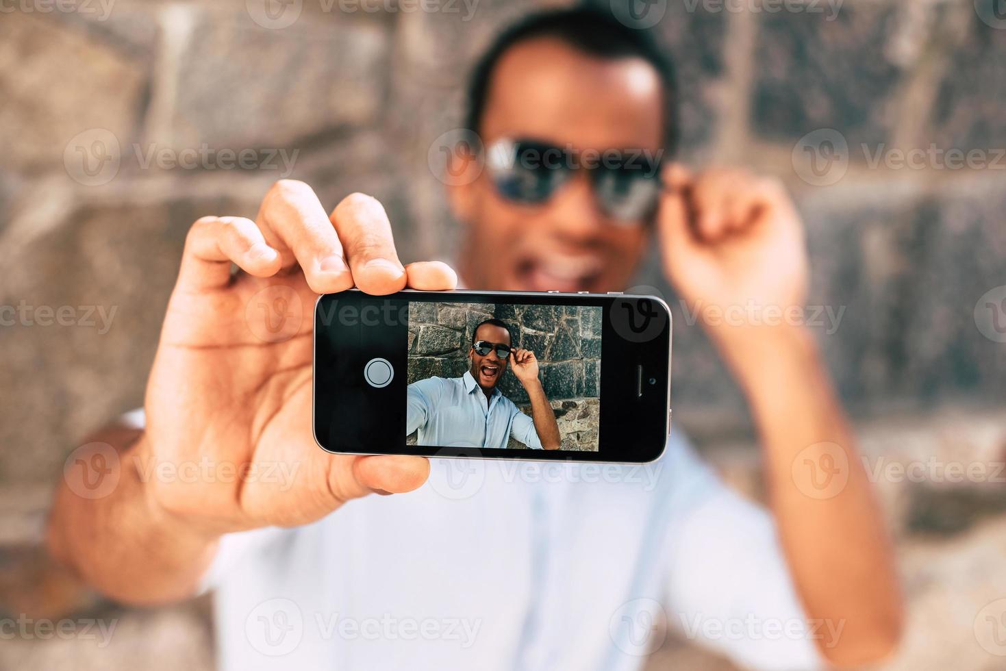 diversão selfie. jovem africano bonito fazendo selfie com seu telefone inteligente e parecendo feliz em pé contra a parede apedrejada ao ar livre foto