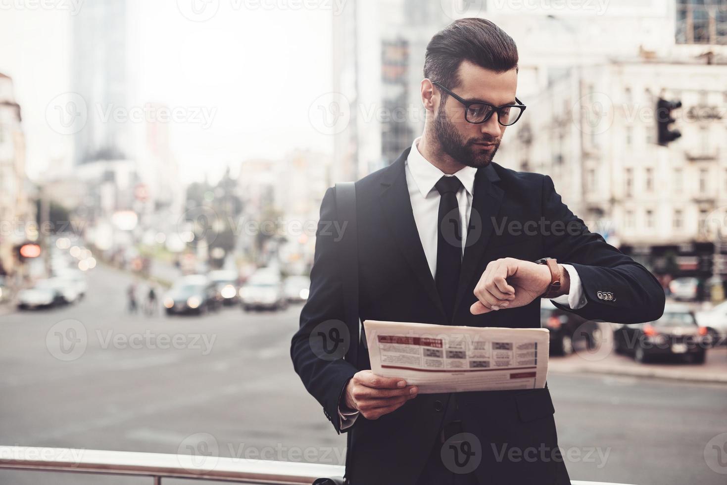 eu deveria estar lá a tempo. jovem confiante de terno completo segurando o jornal e olhando para o relógio em pé ao ar livre com a paisagem urbana ao fundo foto