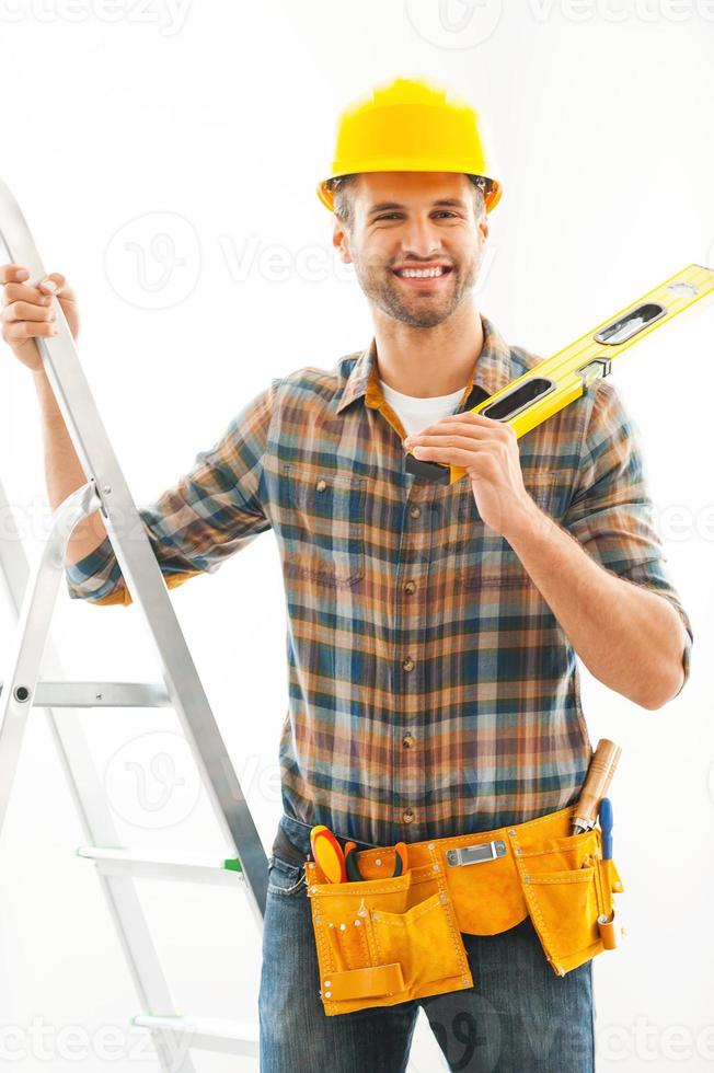 bonito e prático. alegre jovem trabalhador manual segurando a mão na escada e carregando o nível no ombro em pé dentro de casa foto