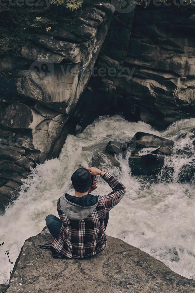 seus melhores tiros. vista superior do jovem moderno fotografando enquanto está sentado na rocha com o rio abaixo foto