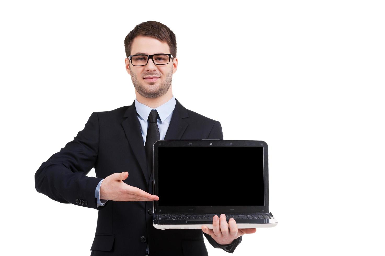 apresentando seu produto. jovem alegre em trajes formais apontando o monitor do computador e sorrindo em pé isolado no branco foto