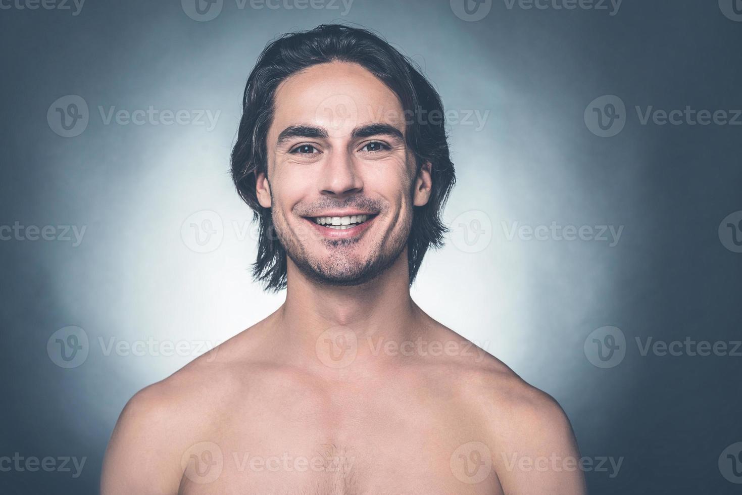 parecendo bem e sentindo-se bem. retrato de jovem sem camisa olhando para a câmera e sorrindo em pé contra um fundo cinza foto