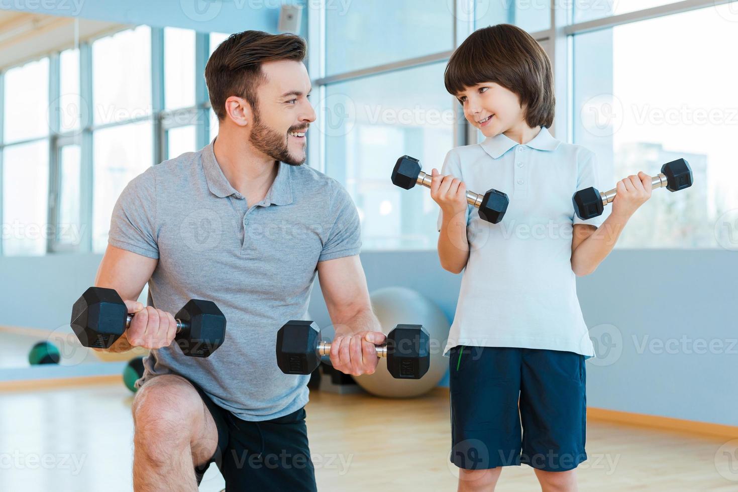 exercitando juntos. feliz pai e filho se exercitando com halteres e sorrindo em pé no health club foto