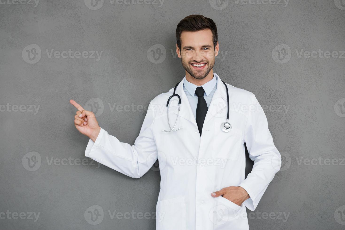 mostrando novas formas de cura. sorrindo jovem médico de uniforme branco, olhando para a câmera e apontando para fora em pé contra um fundo cinza foto