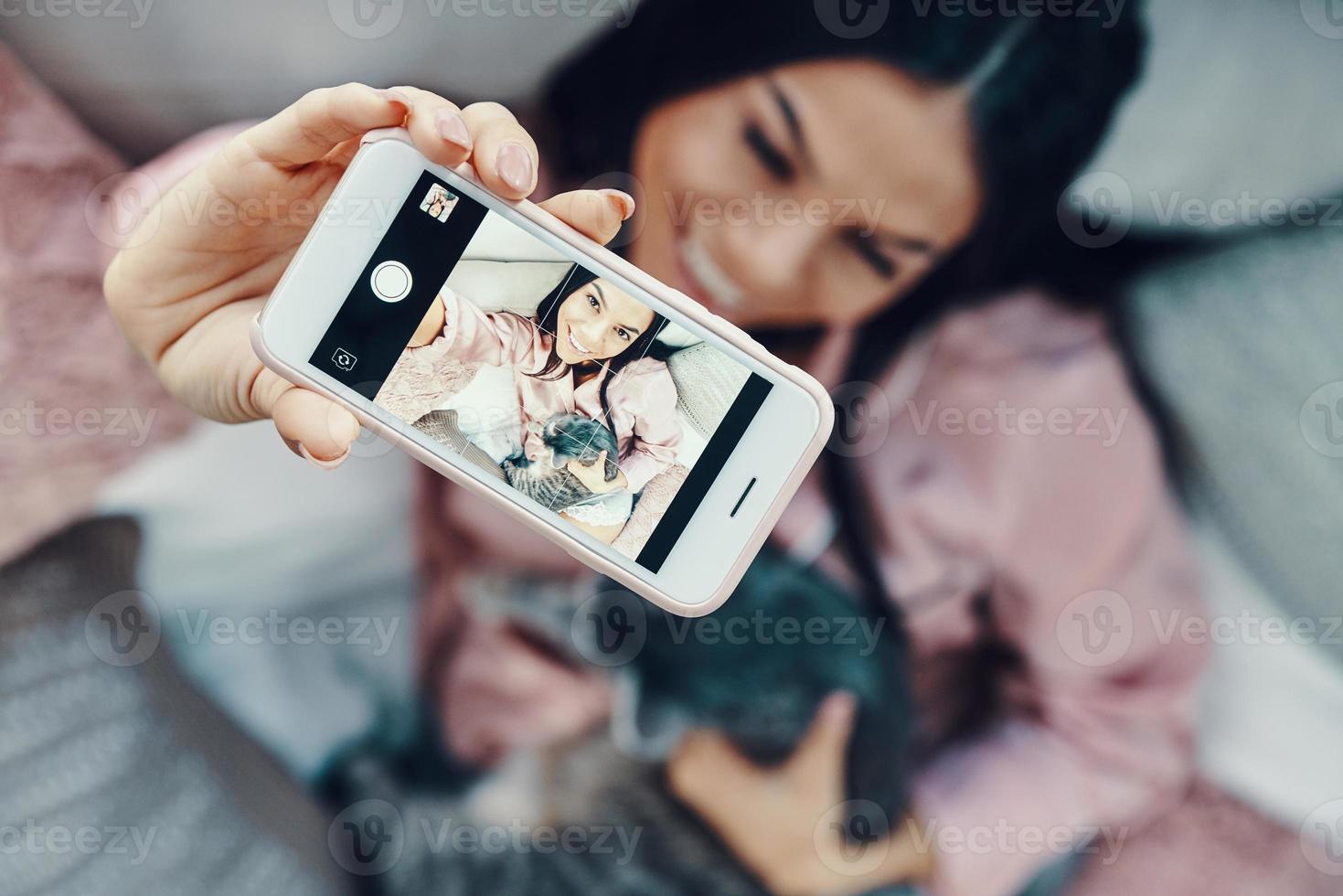 vista superior da bela jovem de pijama abraçando gato doméstico e tomando selfie enquanto descansava na cama em casa foto