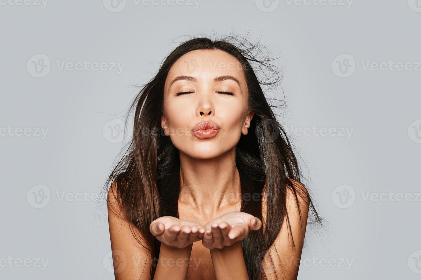 enviando amor. bela jovem mulher asiática, mantendo os olhos fechados e mandando um beijo em pé contra um fundo cinza foto