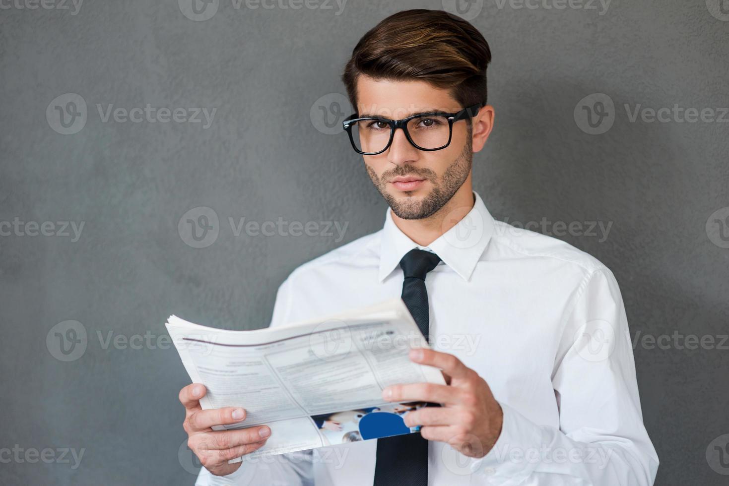 lendo algumas notícias de negócios. jovem confiante de camisa e gravata segurando jornal e olhando para a câmera em pé contra um fundo cinza foto