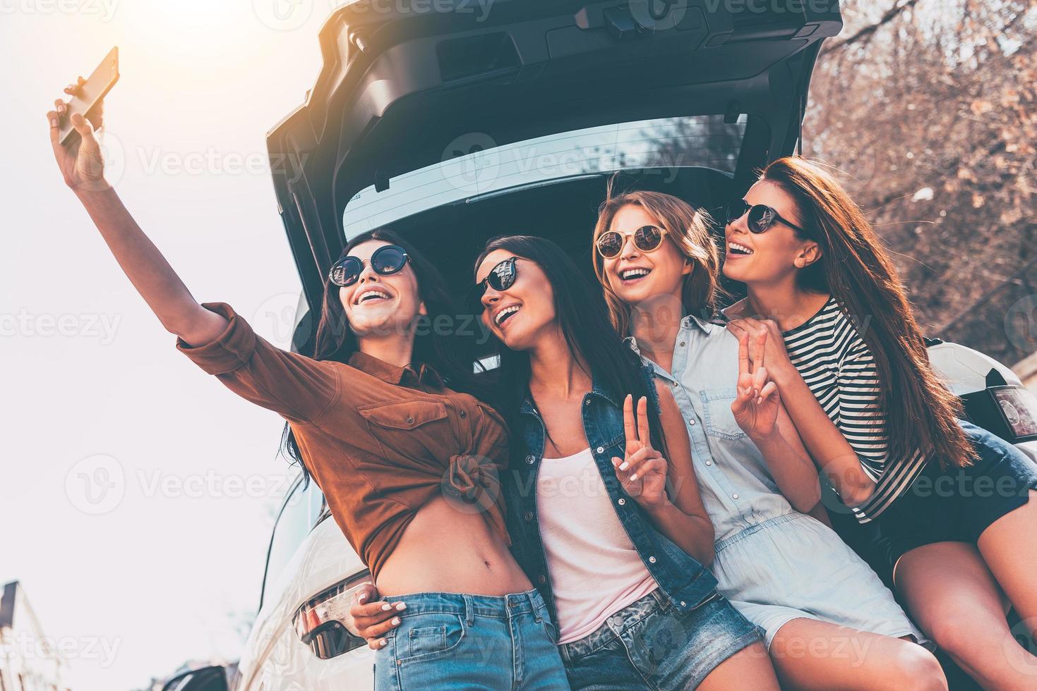 apenas uma selfie antes da viagem quatro belas jovens alegres fazendo selfie com sorriso em pé perto do porta-malas do carro foto