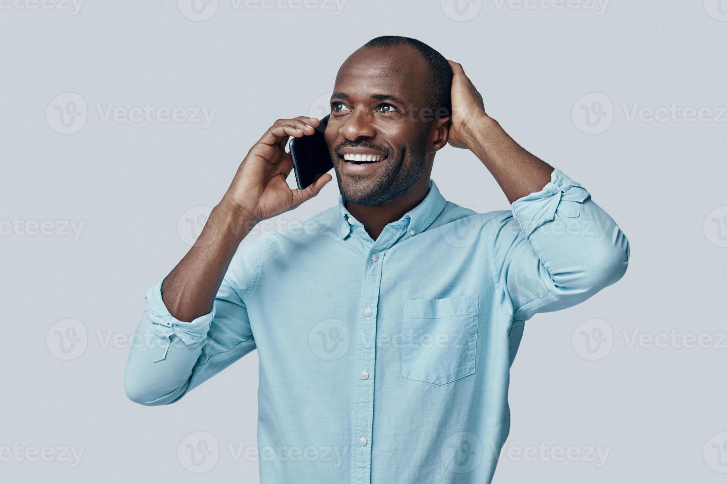 feliz jovem africano falando no telefone inteligente e sorrindo em pé contra um fundo cinza foto