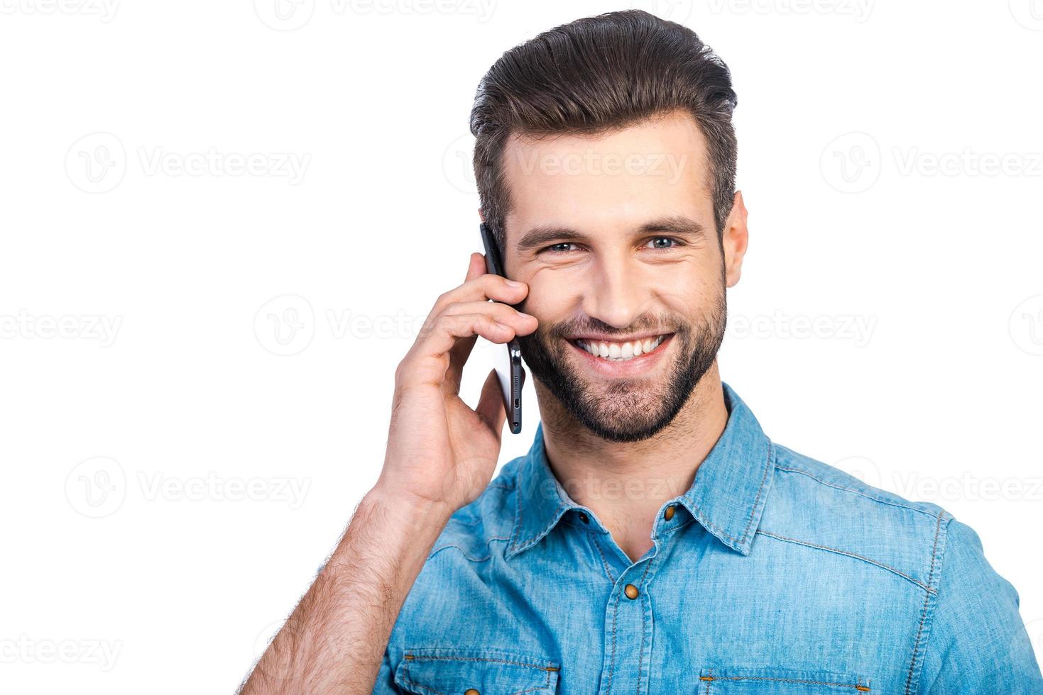 olá confiante jovem bonito na camisa jeans falando no celular e sorrindo em pé contra um fundo branco foto