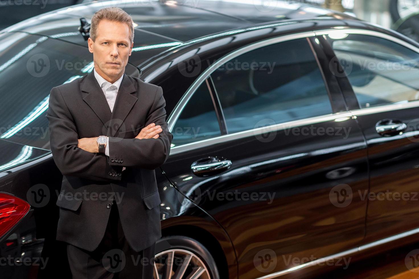 eu amo carros de luxo. vista superior do homem de cabelo grisalho confiante em traje formal, inclinando-se para o carro e olhando para a câmera foto