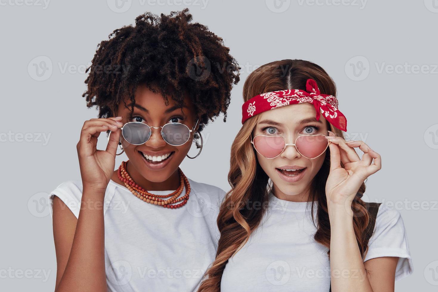 duas mulheres jovens surpresas olhando para a câmera e sorrindo em pé contra um fundo cinza foto