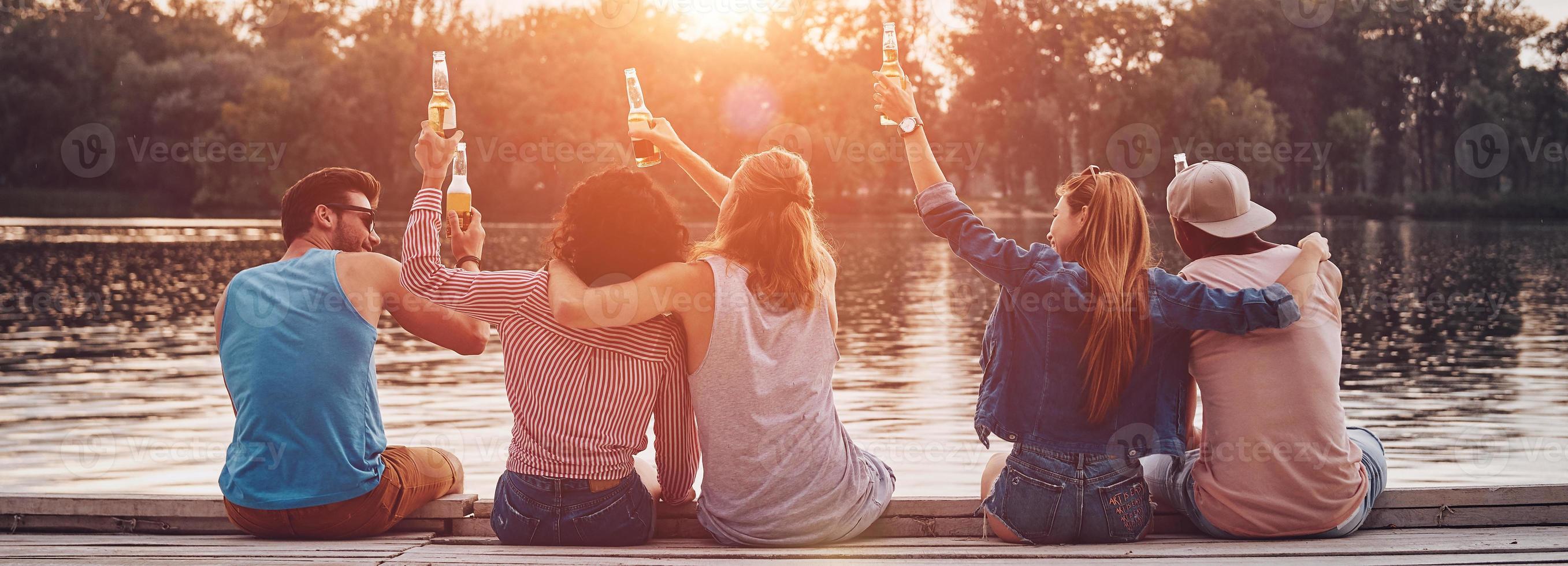 vista traseira de jovens em roupas casuais, brindando com garrafas de cerveja enquanto está sentado no cais foto