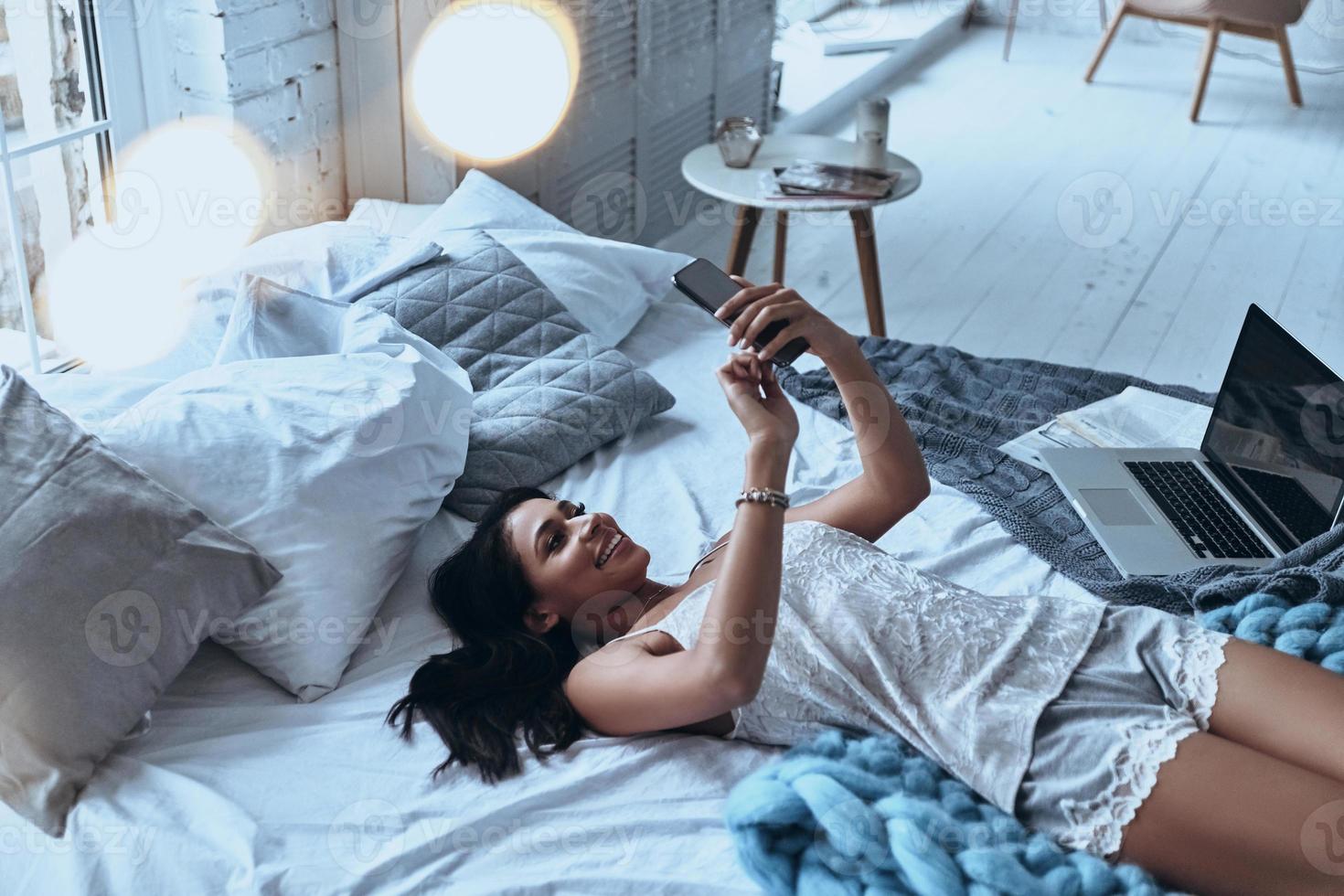 manhã começa com um bom bate-papo. vista superior de uma jovem atraente usando telefone inteligente e sorrindo enquanto estava deitado na cama em casa foto